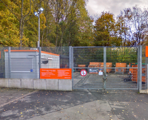 Eingezäunt stehen mehrere orangene Container, im Hintergrund sind Bäume