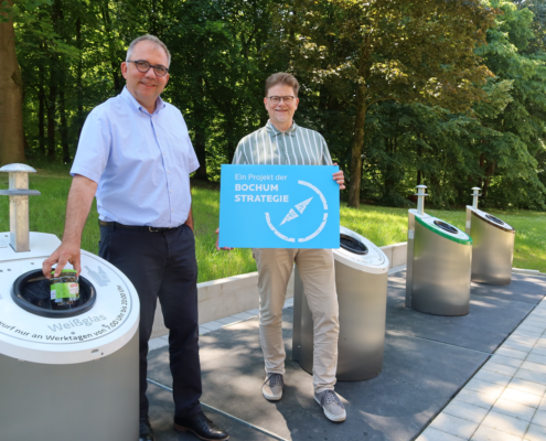 Ein Mann entsorgt Weißglas. Neben ihm steht ein weiterer Mann mit einem Schild mit Aufschrift Ein Projekt der Bochum Strategie