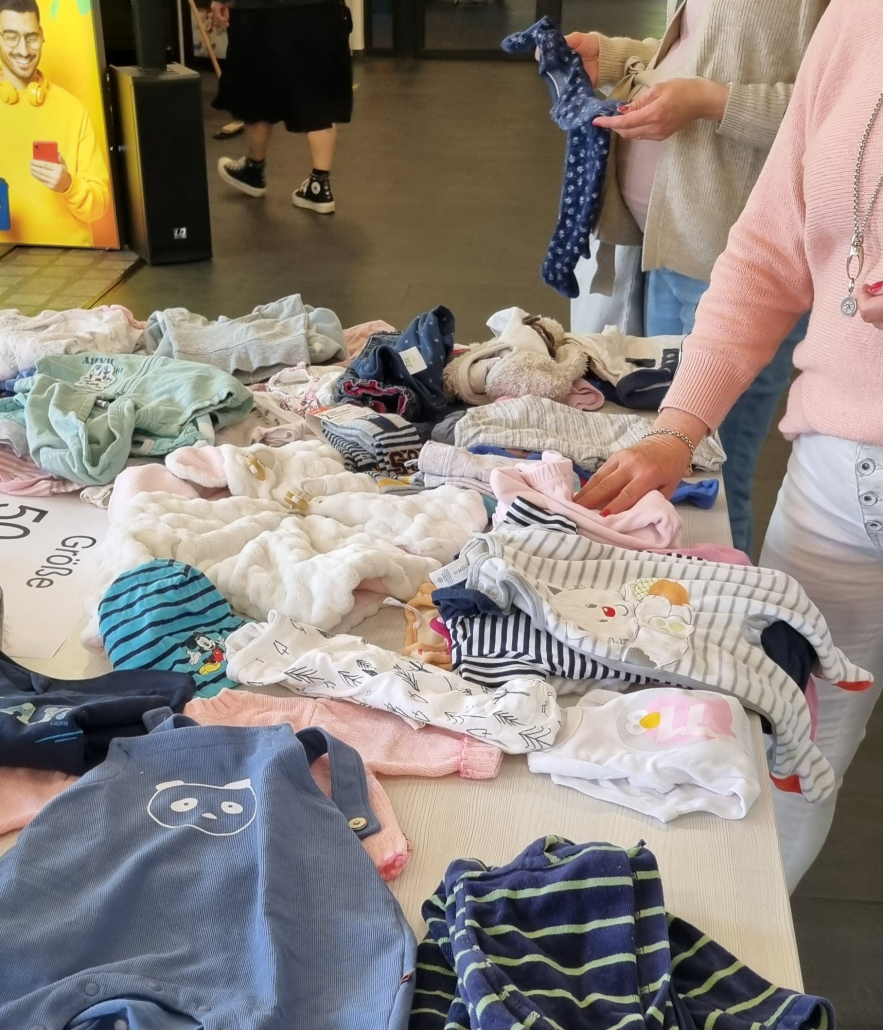 Auf einem Tisch liegen Babyklamotten zwei Frauen schauen sich die Sachen an