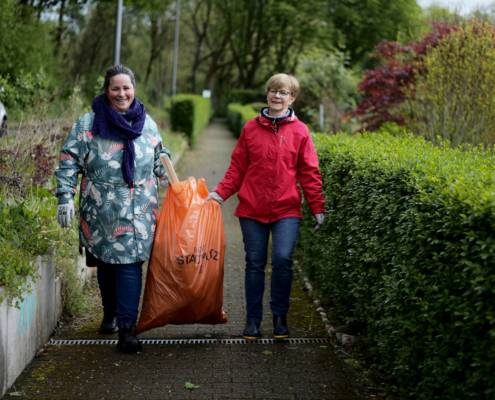 Zwei Frauen tragen einen orangenen Müllsack. sie befinden Sich auf einem Gehweg umrandet mit Büschen.