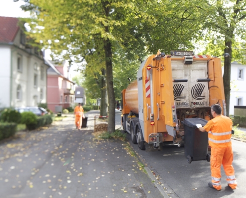 Ein Mitarbeiter in orangener Arbeitskleidung hängt eine Tonne an den Müllwagen, im Hintergrund zieh ein anderer eine Tonne zum Müllwagen.