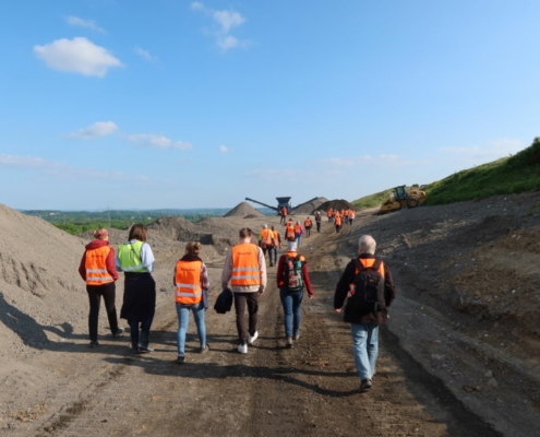 baustellenfuehrungen Eine Gruppe von Menschen schaut sich eine Baustelle an. Im Hintergrund ist eine grüne Landschaft und ein Bagger zu sehen