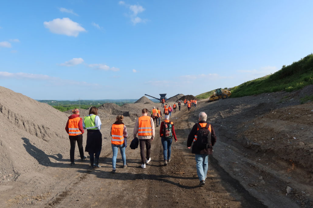 baustellenfuehrungen Eine Gruppe von Menschen schaut sich eine Baustelle an. Im Hintergrund ist eine grüne Landschaft und ein Bagger zu sehen